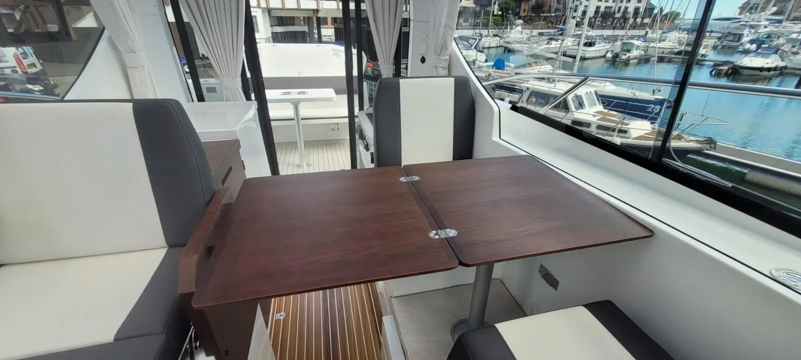 Interior of a modern boat with a wooden table, seating area, and large windows displaying a marina with moored boats.